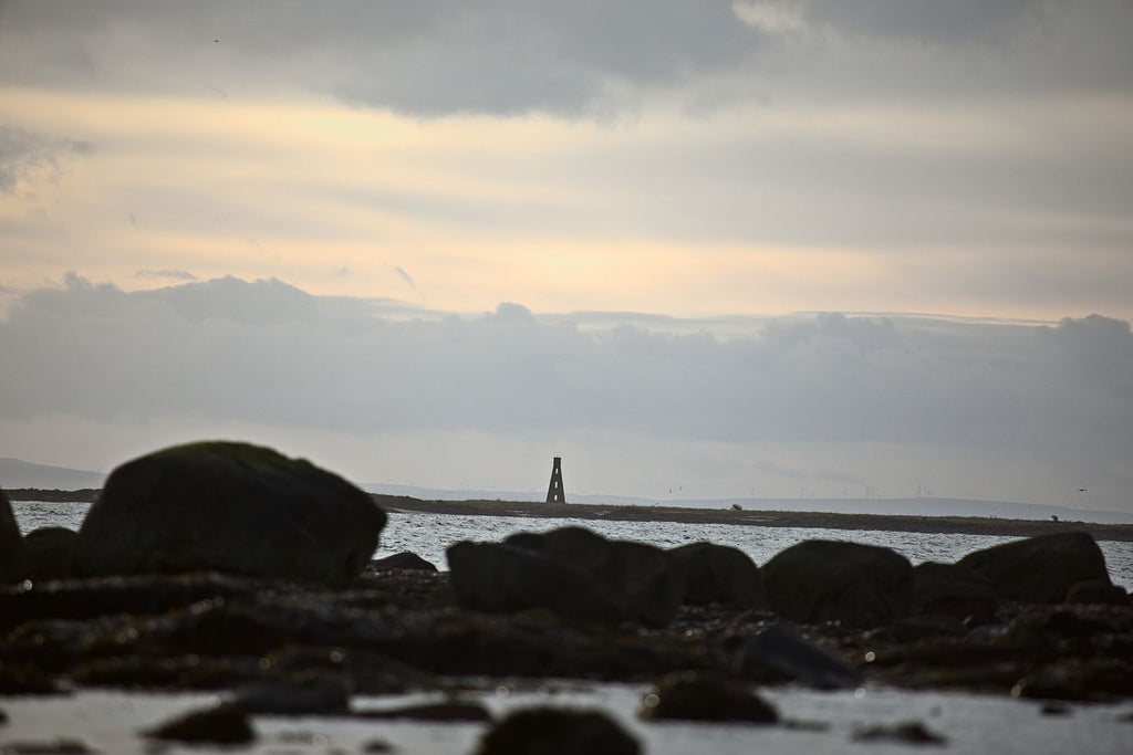 A distant beacon tower beyond dark boulders on a low-tide shore, under a soft evening sky