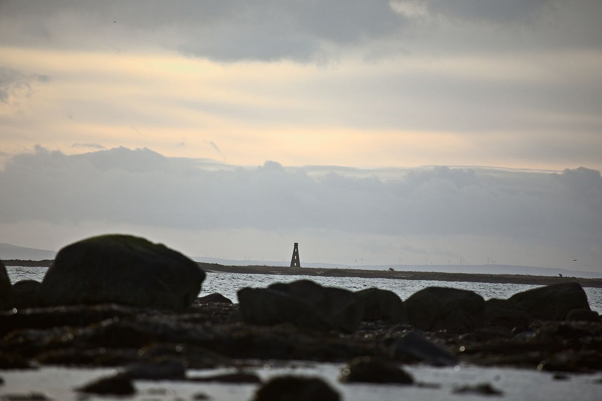 A distant beacon tower beyond dark boulders on a low-tide shore, under a soft evening sky