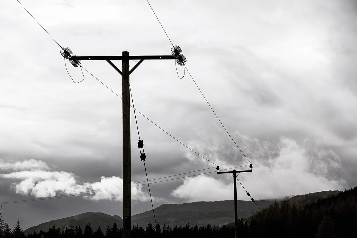 Telegraph poles and wires against a dramatic cloudy sky above dark hills, in black and white