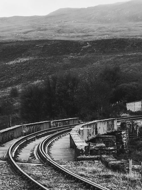 A railway line curving over a stone bridge into dark moorland hills, Scottish Highlands, in black and white