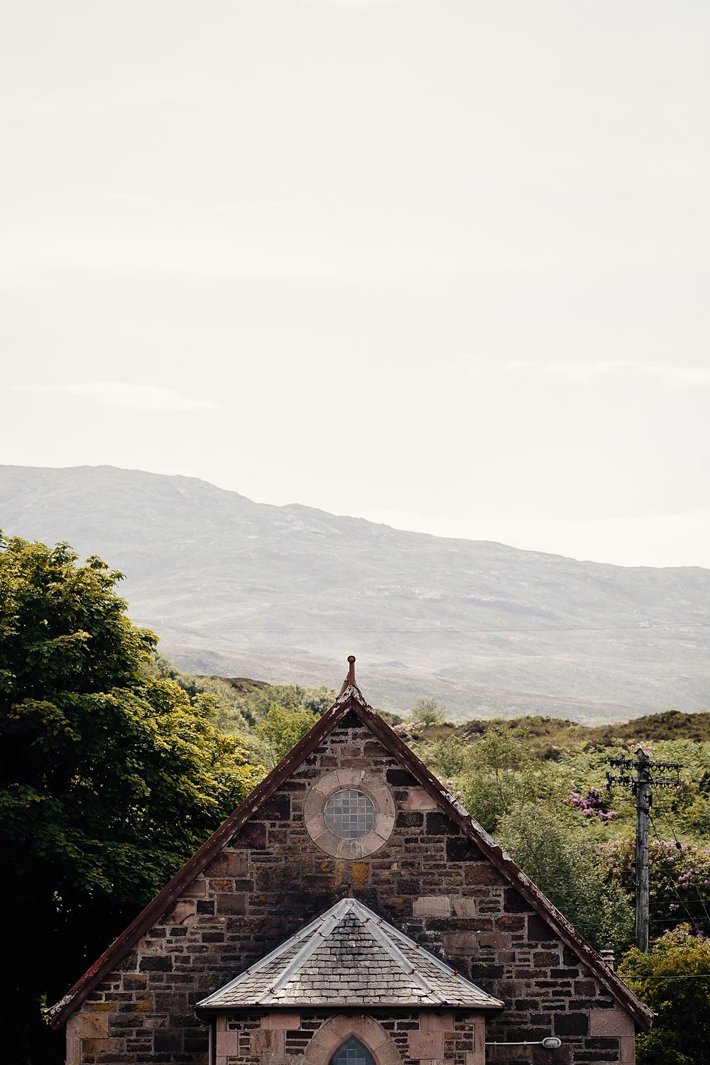 The stone gable of a Highland chapel with a round window, hills and trees behind