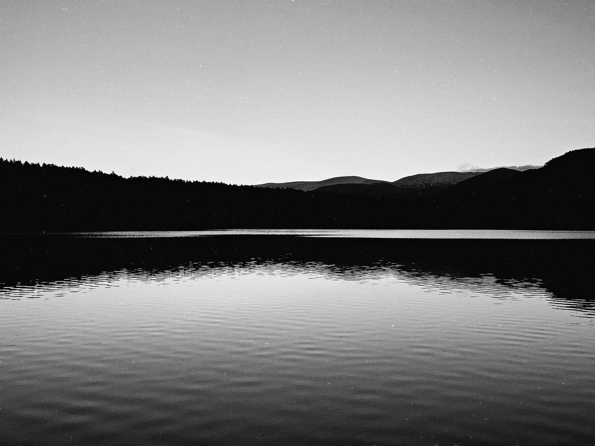 A still loch below a dark tree line and distant hills in the Cairngorms, in black and white