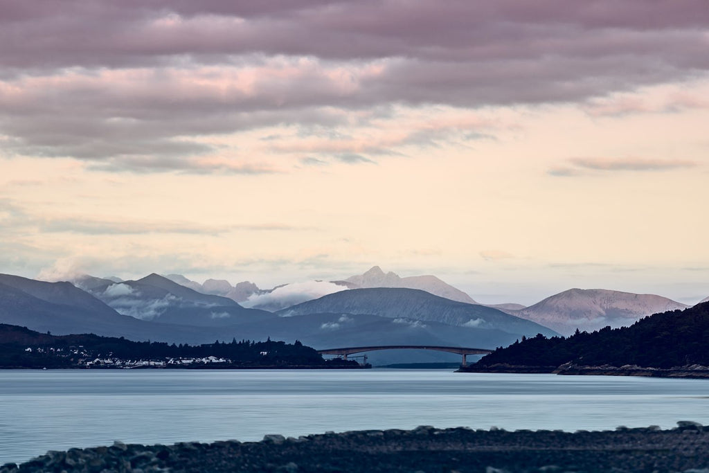 The Skye Bridge below snow-dusted mountains at dusk, under a pink and violet sky