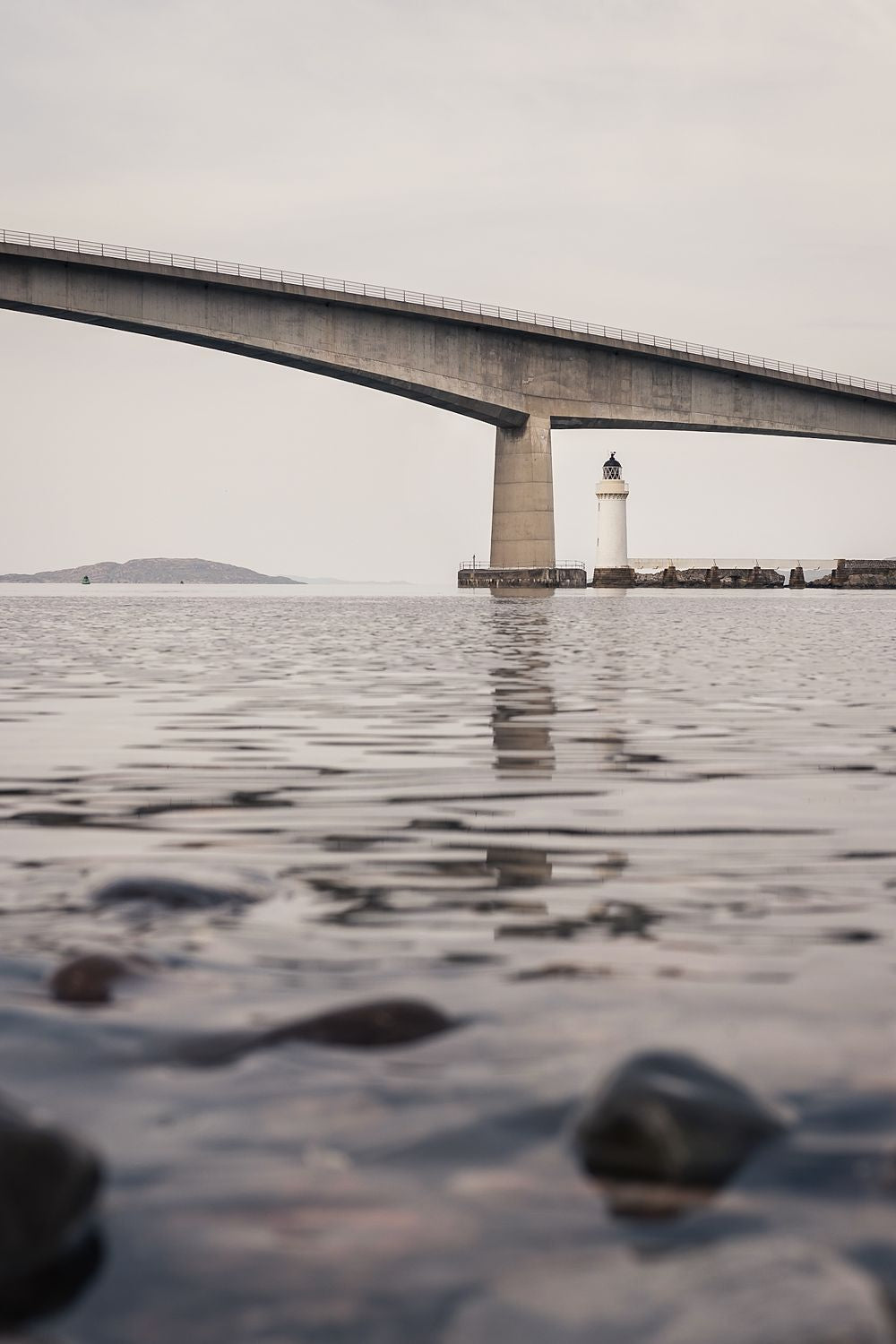 The Skye Bridge and Kyleakin lighthouse seen from the water's edge, ripples and stones in front