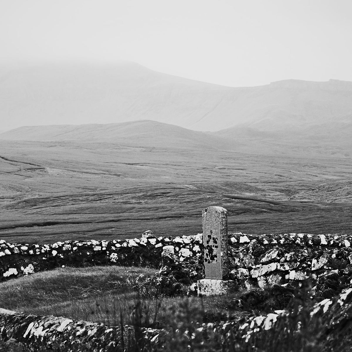 A lichen-covered standing stone by a drystone wall below misty moorland hills, in black and white