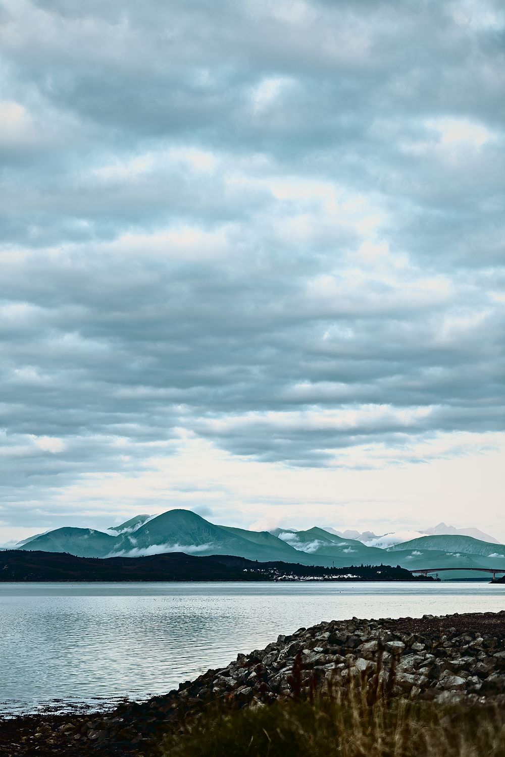 Snow-dusted mountains across a still Highland loch under a big cloudy sky, with a rocky shore in front