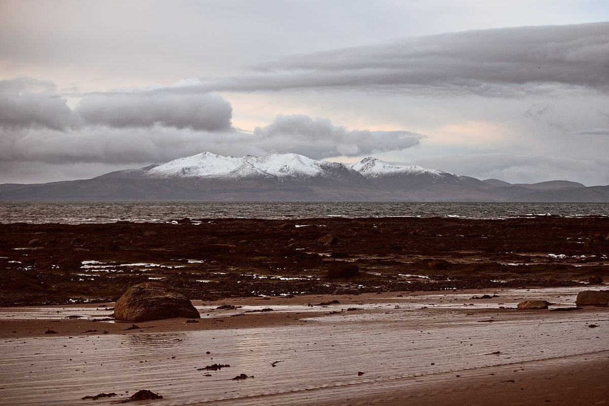Snow-capped hills across the sea beyond a rocky beach at low tide, under a mauve sky