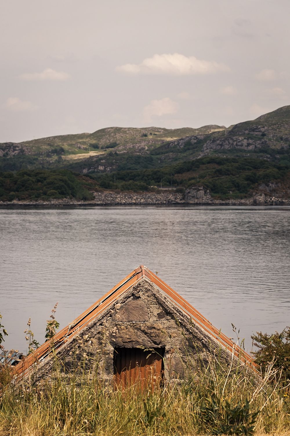 The slate roof of an old stone shed in long grass above a loch, hills beyond