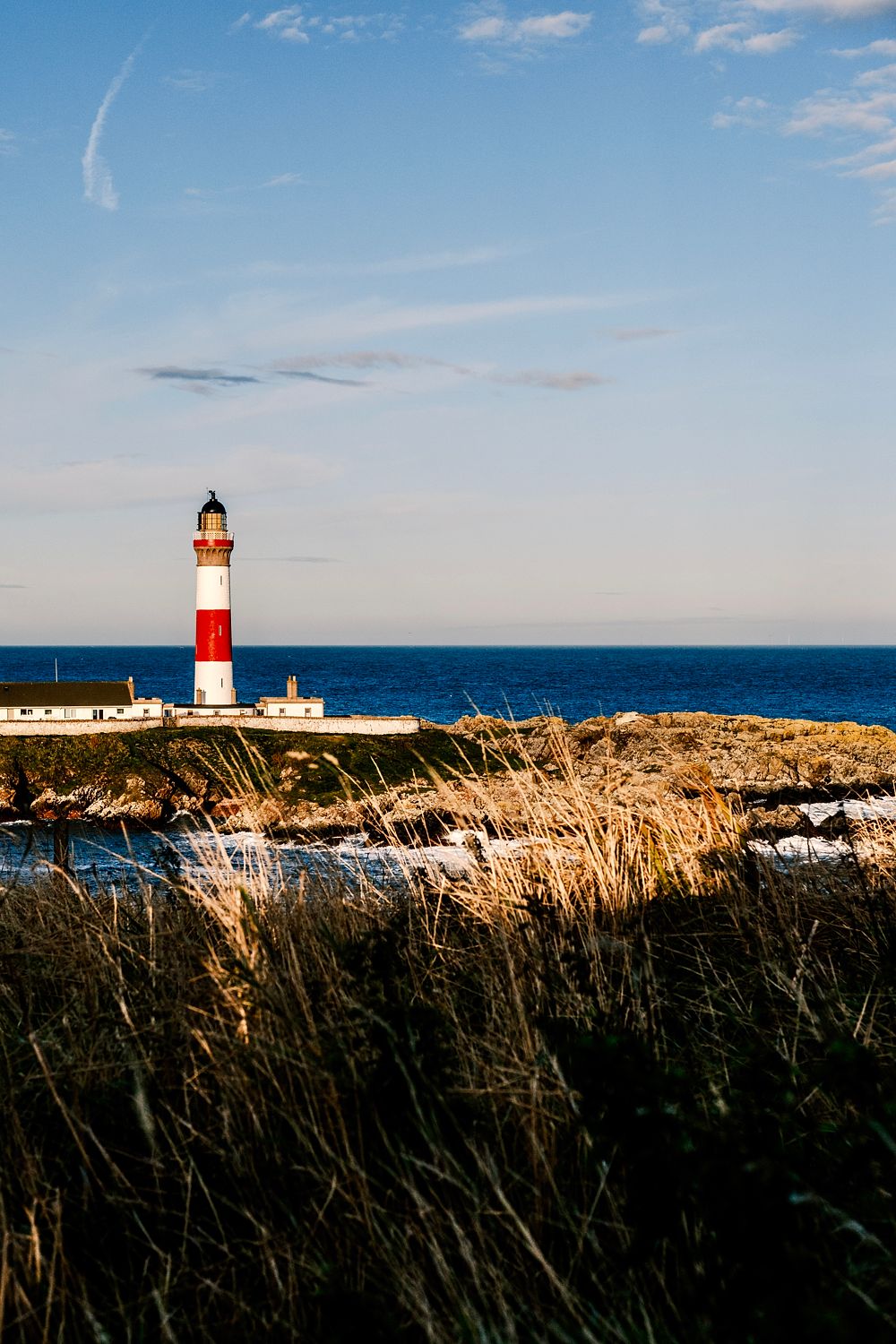 Red and white lighthouse beyond wind-blown coastal grasses and a deep blue sea, Aberdeenshire