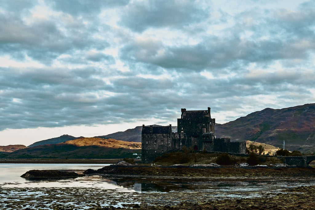 Eilean Donan Castle on its island at low tide, with sunlit hills and a cloudy sky, Scottish Highlands