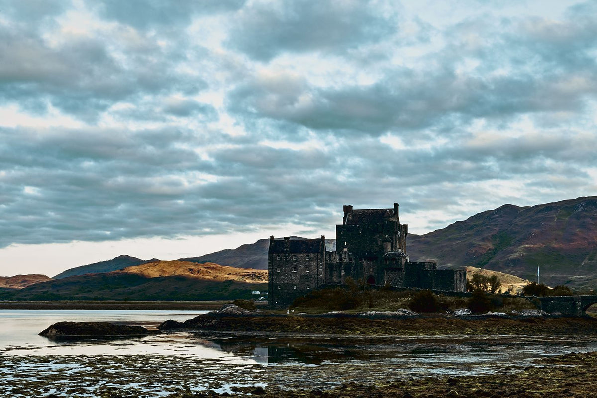 Eilean Donan Castle on its island at low tide, with sunlit hills and a cloudy sky, Scottish Highlands