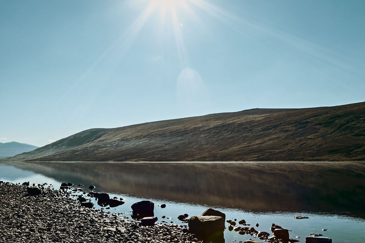 The sun bursting over a hillside above a still loch and pebble shore, Scottish Highlands